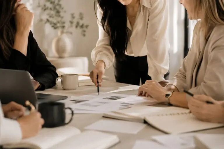 Vier Frauen bei einer Team-Besprechung am Tisch: eine steht und zeigt mit einem Stift auf ein ausgelegtes Layout, während die anderen mit Laptop, Notizbüchern und Kaffeetassen zuhören
